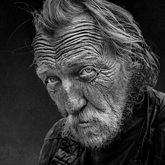 A black and white photo of an old man with very wrinkly skin, a scruffy beard and unkempt hair.