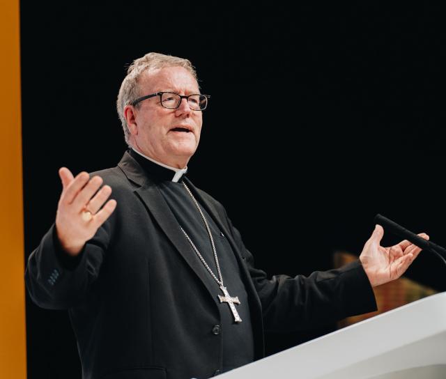 Bishop Robert Barron in priestly garb standing at a pulput with hands outstretched.