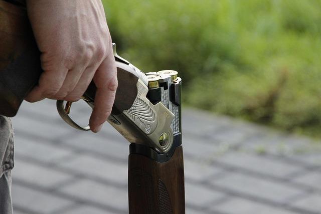 Close up of a man's hand holding a shotgun down at his side, barrel pointing toward the ground.