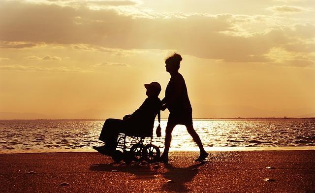 The silhouette of a woman pushing a man in a wheelchair along a beach at sunset.