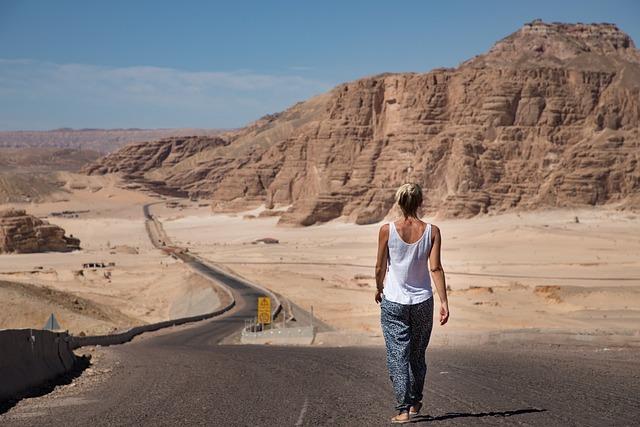 A woman walking alone along a desert road toward rocky hills.