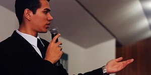 A man with dark hair is holding a microphone and his left hand is held out, palm up as he speaks.
