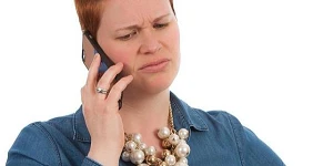 A woman with short red hair and a blue top on the phone looking puzzled.